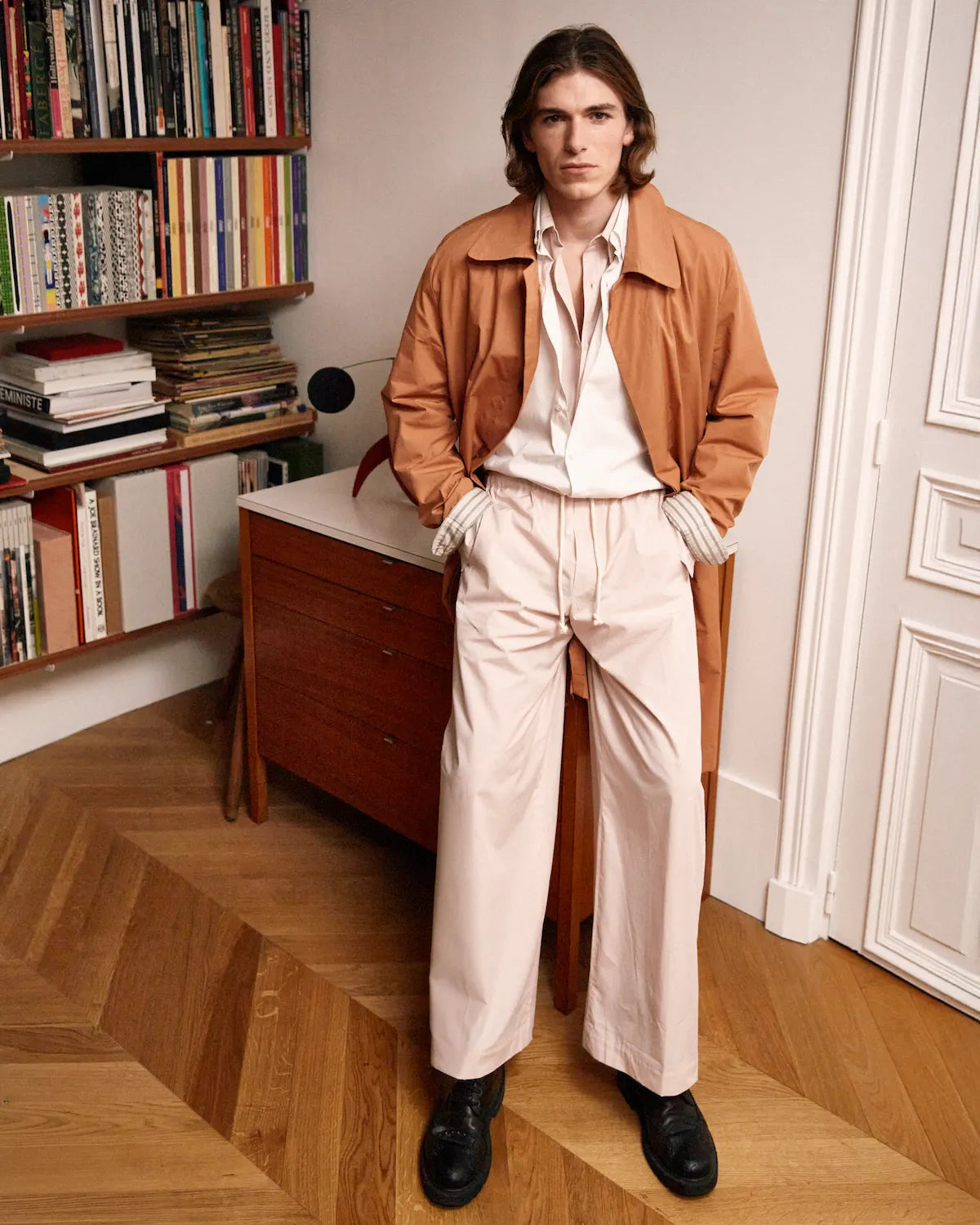 homme portant un manteau terracotta et un ensemble chemise pantalon rose meus paris dans une bibliothèque