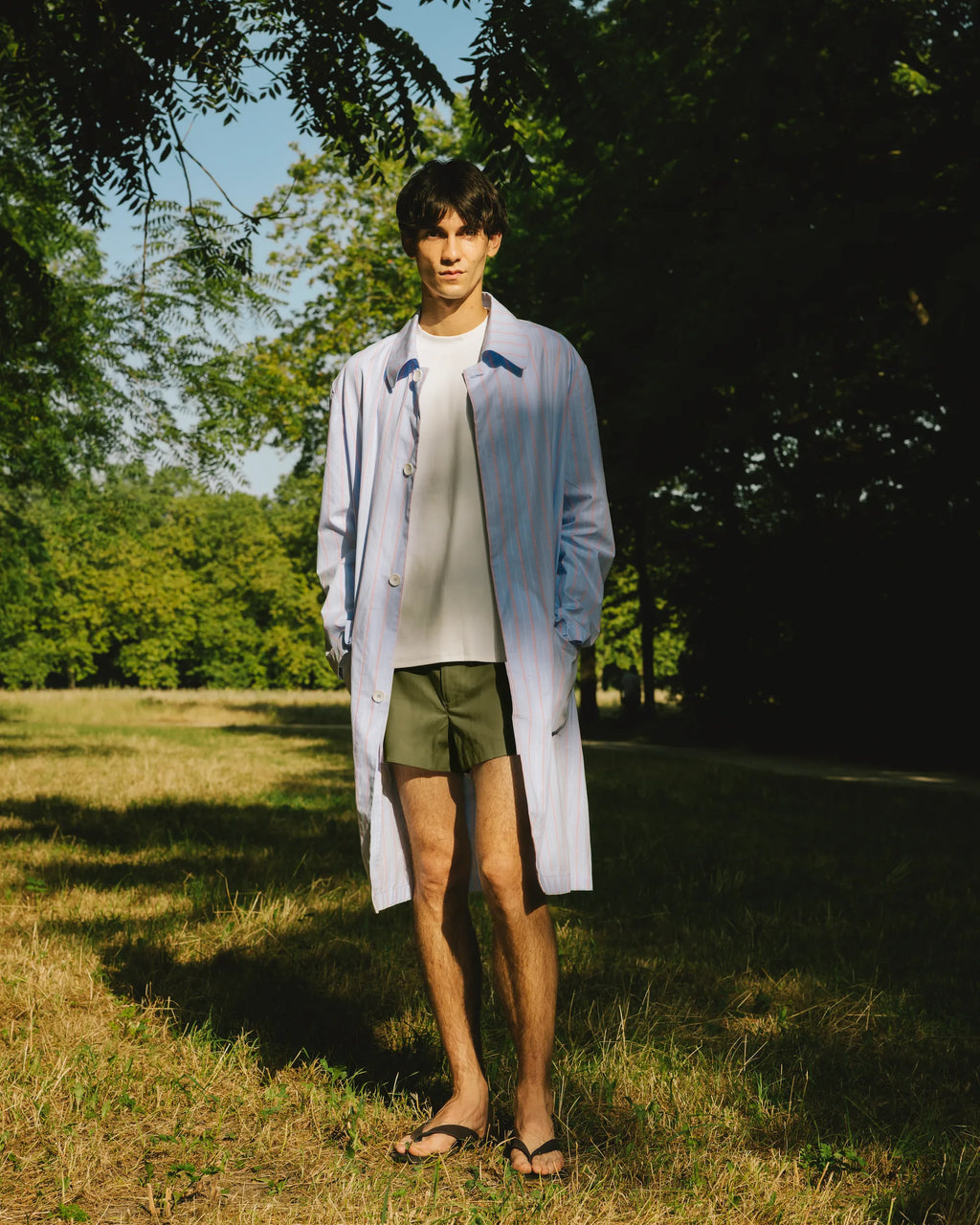 homme portant un manteau léger en coton bleu rayé rouge et un short kaki meus paris dans un bois 